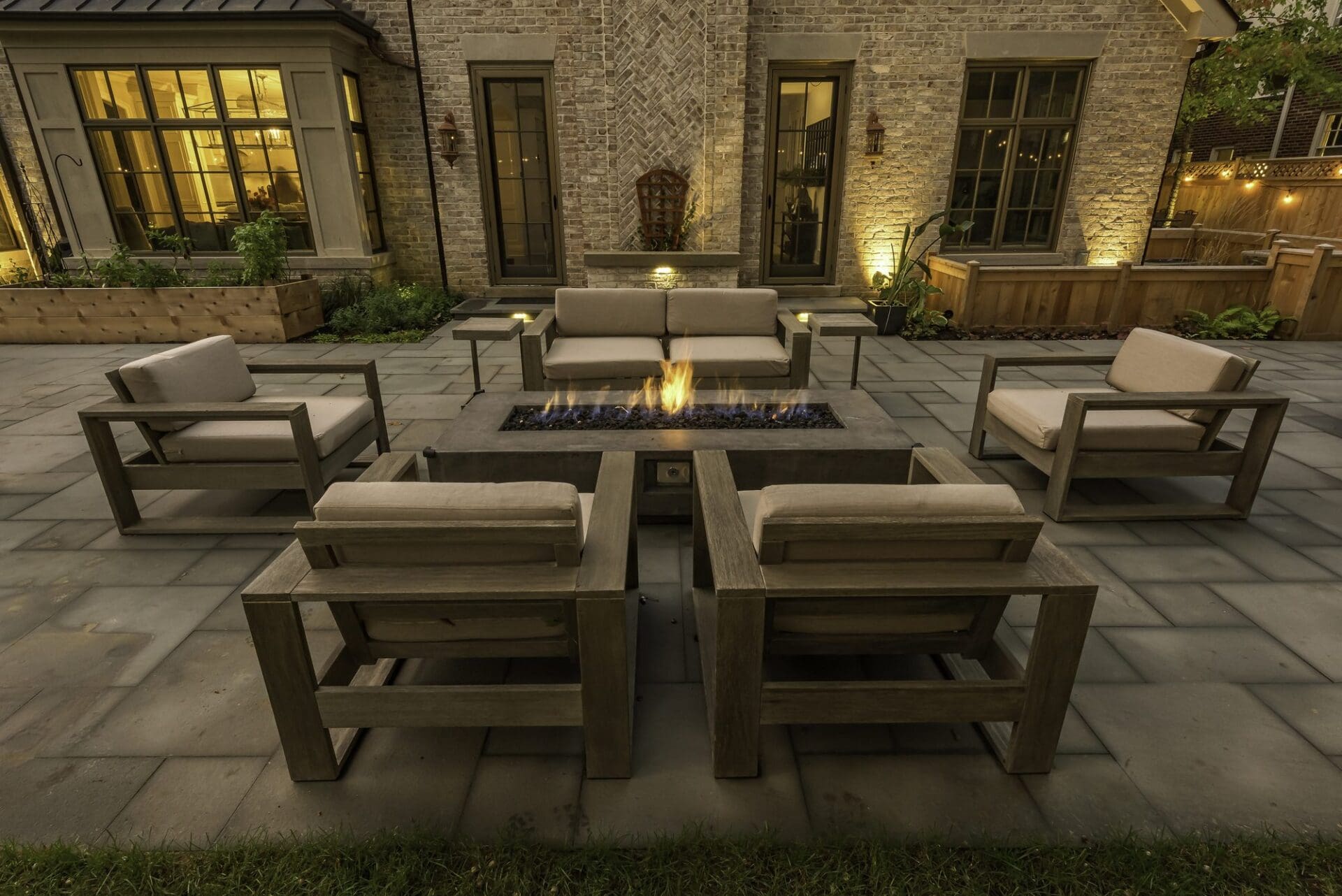 Backyard paver patio at dusk with lit fire tables and lounge seating — Apex Landscaping